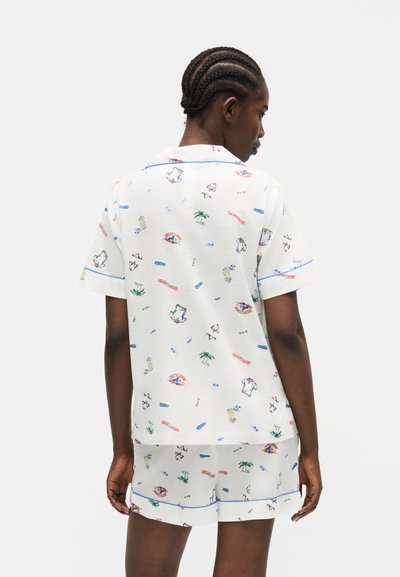 Άτομο που φοράει λευκό κοντομάνικο πουκάμισο και σορτς με πολύχρωμα μοτίβα ταξιδιωτικού θέματος, στέκεται με την πλάτη προς την κάμερα.