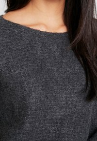 Pull en maille grise avec un large décolleté et une texture douce, présentant un ourlet légèrement irrégulier aux épaules et un gros plan sur le tissu.