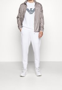 Grå lätt jacka, vit sweatshirt med en marinblå logotyp, vita sweatpants och grå sneakers. Släta ytor och avslappnad passform.