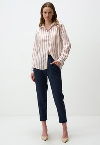 Camicia bianca a maniche lunghe con strisce verticali rosse, chiusura a bottoni sul davanti e polsini arrotolati; abbinata a pantaloni blu scuri a sigaretta e tacchi color beige.