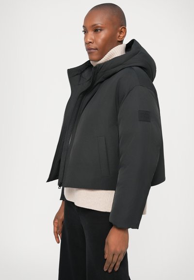 Μαύρο κοντό παλτό με κουκούλα, με φερμουάρ, πλαϊνές τσέπες και μια υφή μαύρη πατσαβούρα στον ώμο.