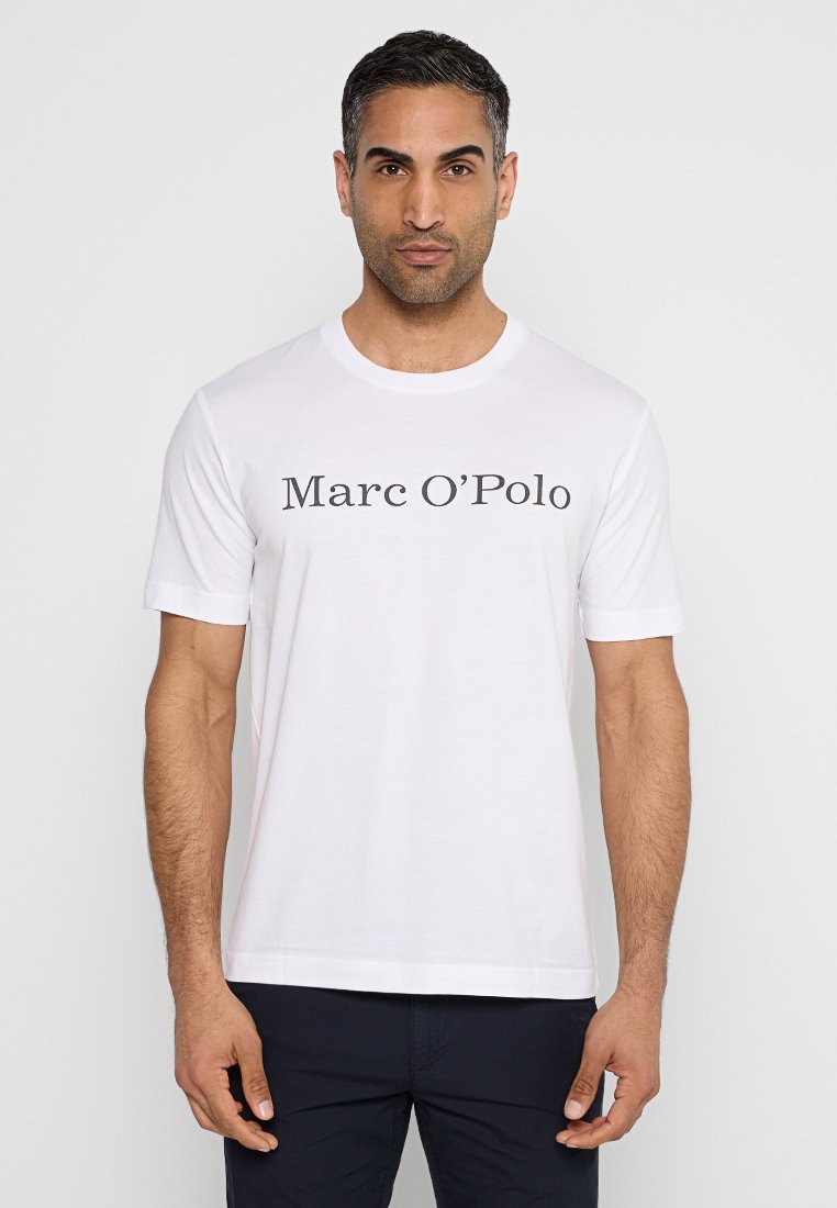 Mann in einem weißen Rundhals-T-Shirt mit dem Text "Marc O’Polo", stehend vor einem hellen, einfarbigen Hintergrund.