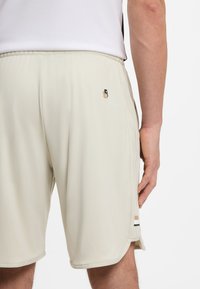 Világos bézs atlétikai rövidnadrág sima anyaggal, apró logóval a hátoldalon és csíkos díszítéssel az oldalakon.