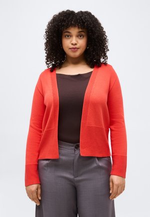 Woman with curly hair wearing a red open-front cardigan, dark brown top, and gray high-waisted pants against a plain background.