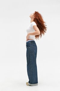 Νεαρή γυναίκα με μακριά σγουρά κόκκινα μαλλιά, λευκό εφαρμοστό t-shirt, φαρδιά μπλε τζιν και εσπαντρίγιες στέκεται σε προφίλ με το κεφάλι της γερμένο πίσω.