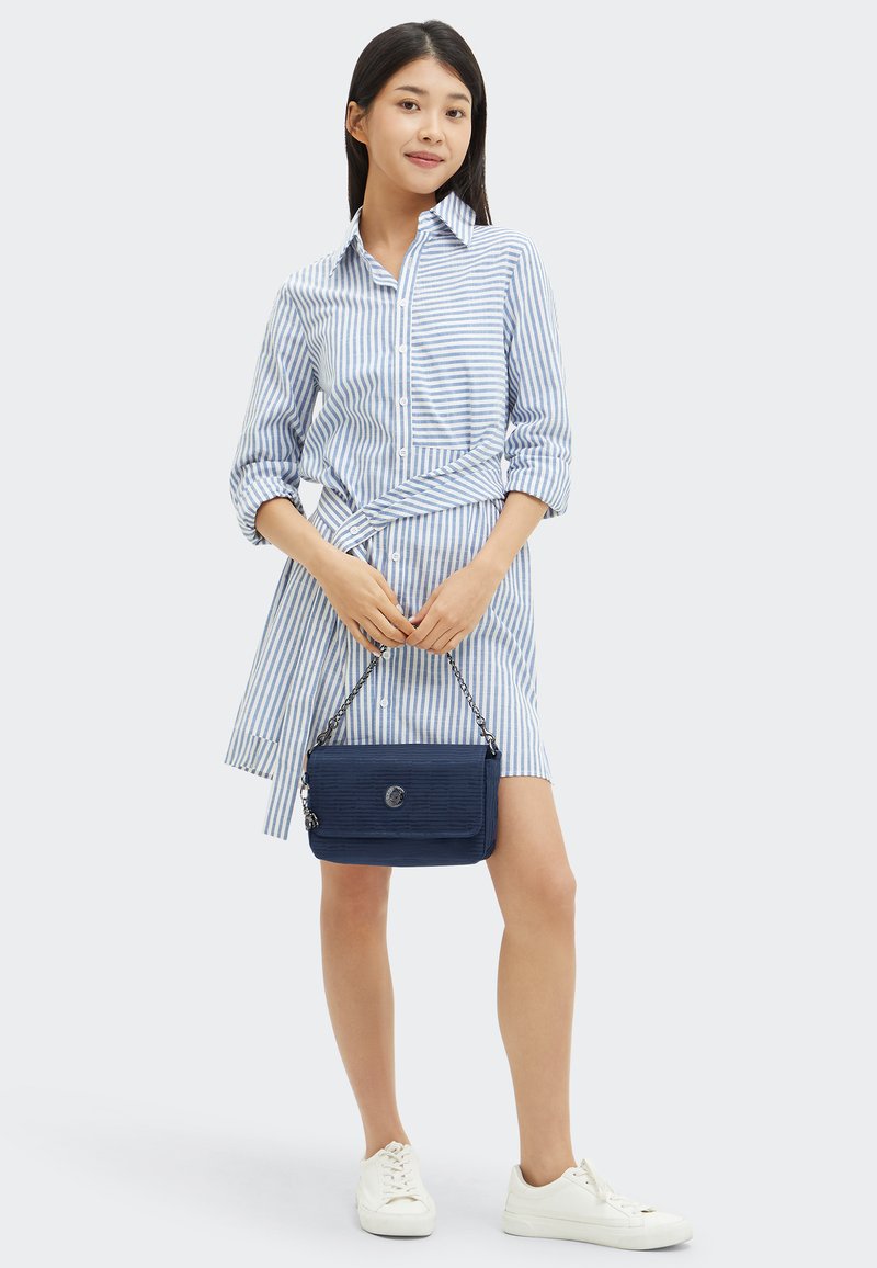 Giovane donna che indossa un vestito camicia a righe blu e bianche, tiene una borsa blu navy con una tracolla a catena, indossa scarpe da ginnastica bianche.