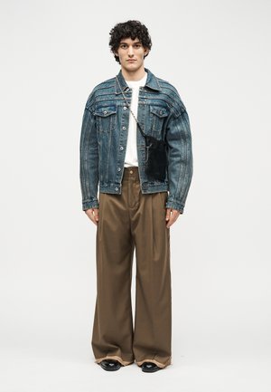 Joven con una chaqueta vaquera azul de gran tamaño, camisa blanca, pantalones anchos marrones, zapatos negros y un pequeño bolso negro de bandolera sobre fondo blanco.