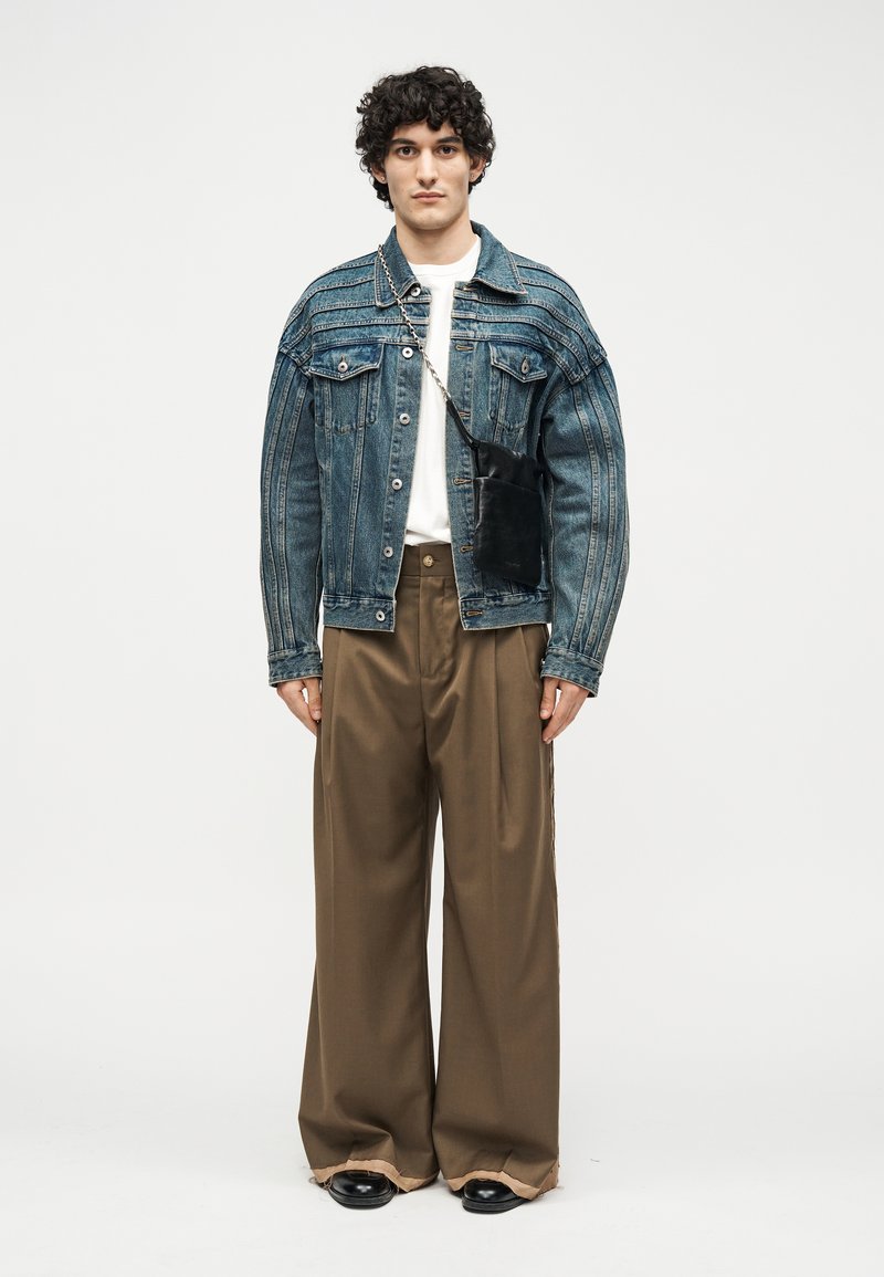 Mladý muž oblečený v nadmernej modrej džínsovej bunde, bielom tričku, širokých hnedom nohaviciach, čiernych topánkach a s malou čiernou kabelkou na ramene na bielom pozadí.