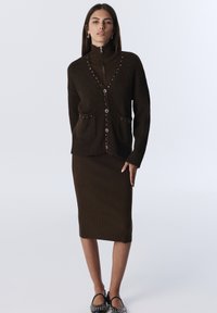 Bruine gebreide cardigan met frontzakken, knoopsluiting en decoratieve knopen. Gecombineerd met een bijpassende ribgebreide bruine rok en zwarte versierde ballerina's.