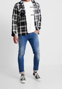 Camicia a scacchi bianca e nera con accenti gialli sopra una maglietta bianca, abbinata a jeans azzurri chiari e sneakers nere.