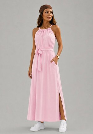 Femme portant une robe maxi rose sans manches avec une fente latérale, un lien à la taille, des baskets blanches, un foulard marron sur la tête et des bijoux dorés, debout devant un fond gris.