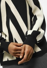 Maglione lavorato a maglia con un motivo a zigzag nero e crema, con polsini a coste e una consistenza morbida. Inquadratura ravvicinata di mani che regolano la manica.