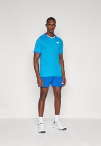 Blue athletic shirt with white collar and logo, paired with blue shorts, worn with white sneakers and crew socks, standing against a plain background.