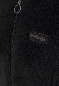 Svart fleecejacka med en mjuk, texturerad yta. Har dragkedja och en etikett med "ONLY PLAY"-märke på framsidan.