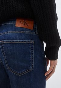 Σκούρο μπλε τζιν παντελόνι με δερμάτινη ετικέτα Calvin Klein, στενή γραμμή, ορατή ραφή και πίσω τσέπη. Μαύρο πλεκτό πουλόβερ με υφή στο χέρι.