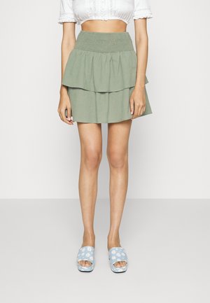 Kobieta ubrana w zieloną, wielowarstwową mini spódniczkę, białą krótką bluzkę i niebieskie klapki z białymi wzorami stokrotek.