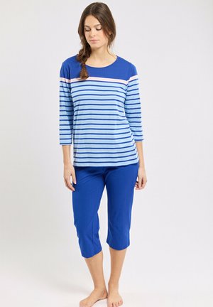 Femme debout pieds nus portant un haut à manches longues rayé bleu et un pantalon court bleu uni sur fond blanc.