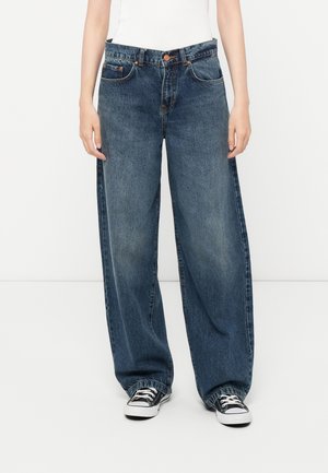 Person wearing high-waist, loose-fit blue jeans with a faded wash and black-and-white sneakers, standing against a plain white background.