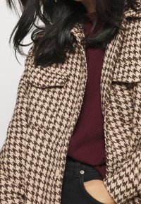 Cappotto a fantasia houndstooth nei toni del marrone e del beige, con due tasche sul petto, chiusura a bottoni e una leggera consistenza, indossato sopra un maglione bordeaux e pantaloni neri.