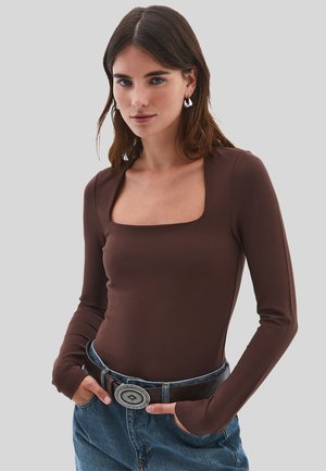 Mujer con cabello castaño liso que lleva pendientes de plata, una blusa marrón de manga larga con escote cuadrado, vaqueros azules y un cinturón con una gran hebilla decorativa.