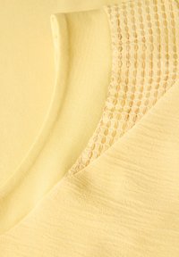 Camicetta gialla con yoke testurizzato a motivo a griglia. Tessuto liscio con una leggera lucentezza, scollatura arrotondata e finitura morbida.