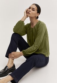 Roheline pikkade varrukatega blouse, millel on volditud detailid, valmistatud siledast kangast. Kuna see on ühendatud tumedate teksadega ja mustade mokassiinidega.