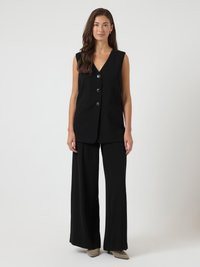 Mulher em pé, vestindo um colete preto sem mangas com botões e calças largas pretas, com sapatos bejes de bico fino, contra um fundo branco.