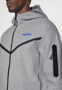 Sudadera con cremallera gris con acentos negros, que presenta un diseño en forma de V, un logo azul y un bolsillo con cremallera en la parte izquierda de la manga. Textura suave.