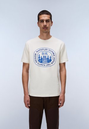 Homme portant un T-shirt blanc avec un graphisme circulaire bleu comprenant des caractères japonais et des arbres, associé à un pantalon marron foncé.