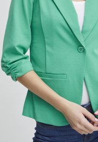 Žalias švarkas, pagamintas iš lygaus, elastingo audinio. Įrengta viena mygtuko uždarymo, sulankstyti rankovės ir priekinė kišenė. Prigludęs dizainas.