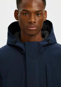 Giacca con cappuccio blu navy dal design liscio, con zip frontale e dettagli di design discreti. Presenta cordini regolabili attorno al cappuccio.