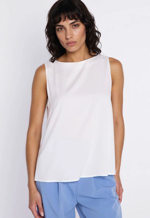 Kobieta o ciemnych kręconych włosach, ubrana w bezrękawnik w kolorze białym i jasnoniebieskie spodnie, na jednolitym białym tle.