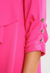 Fuchsia blus med en lätt, slät textur. Har rullade ärmar som säkras med silverknappar och en draperad framdesign.