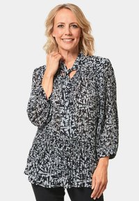 Zwart-witte blouse met een patroon, ronde halslijn en strikdetail, lange geplooide mouwen en een geraffineerde taille. Lichtgewicht, doorschijnende stof.