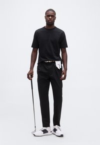 Άνδρας με μαύρη ενδυμασία κρατάει μπαστούνι γκολφ, λευκά γάντια γκολφ κρυμμένα στη ζώνη, στέκεται μπροστά σε απλό φωτεινό φόντο.