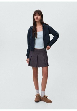 Granatowa bluza z zamkiem, jasnoniebieska bluzka na ramiączkach, plisowana szara spódnica, białe skarpetki i brązowe sneakersy. Modelka stoi z jedną ręką na biodrze.