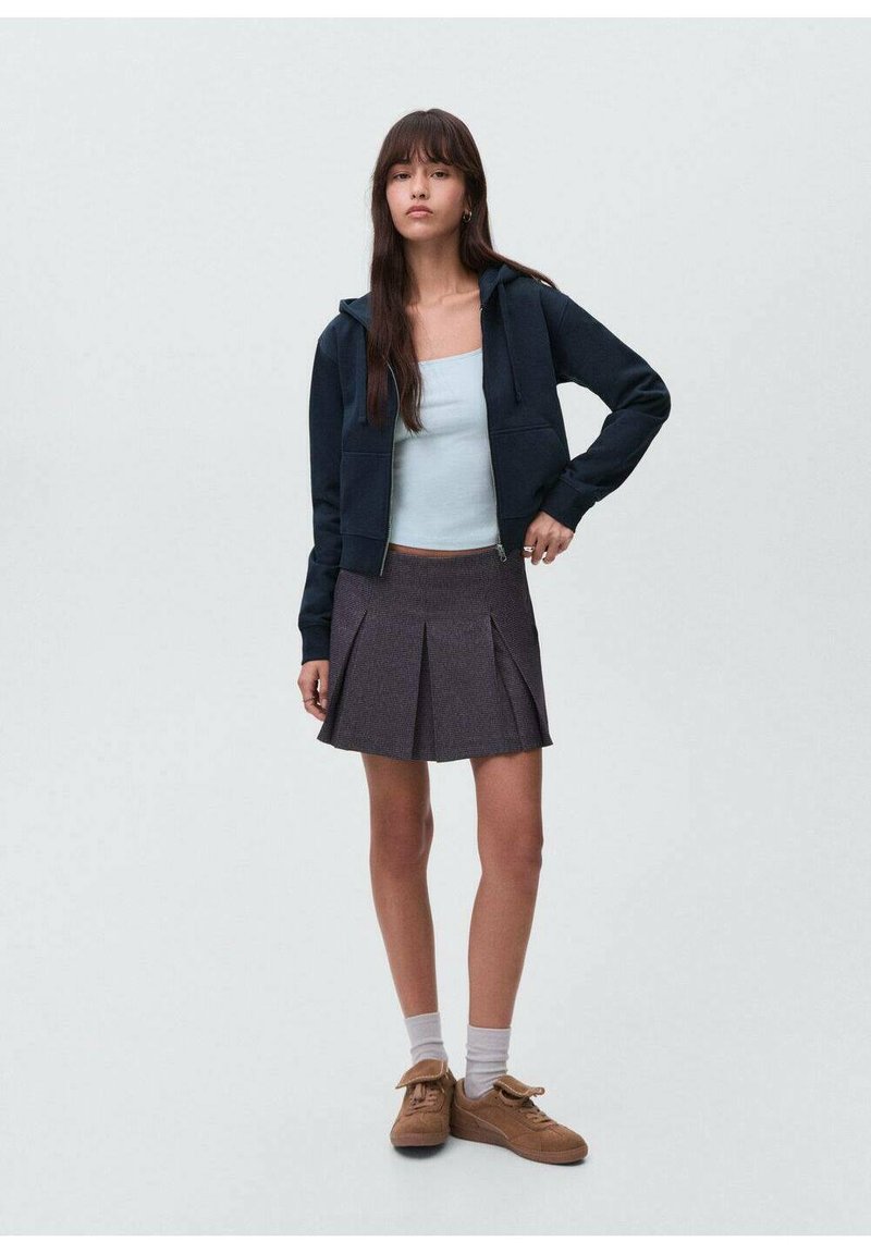 Mornarska zip-up dukserica, svijetloplava majica bez rukava, naborana siva suknja, bijele čarape i smeđi tenisice. Model stoji s jednom rukom na kuku.