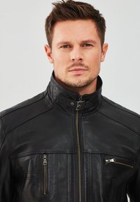 Giubbotto di pelle nera con colletto alto, chiusura con zip frontale, dettagli di cucitura orizzontale e diagonale, e due tasche con zip.