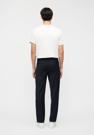 Άνδρας στέκεται πλάγια, φοράει απλό λευκό κοντομάνικο μπλουζάκι, μαύρο ραμμένο παντελόνι και λευκά αθλητικά παπούτσια, σε λευκό φόντο.