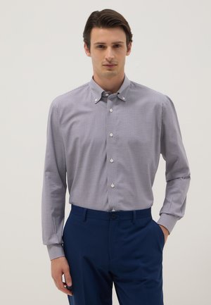 Uomo che indossa una camicia a maniche lunghe con piccoli quadretti, abbottonata e infilata nei pantaloni blu scuro, in piedi con una mano in tasca.