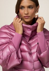 Chaqueta acolchada rosa con un exterior brillante y acolchado, cuello alto y suaves puños acanalados. Cuenta con dos botones en el cuello para el cierre.