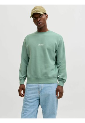 Homme portant un sweat-shirt vert clair avec le texte blanc « JJ Studios », jean bleu clair et casquette de baseball beige, debout devant un fond uni.