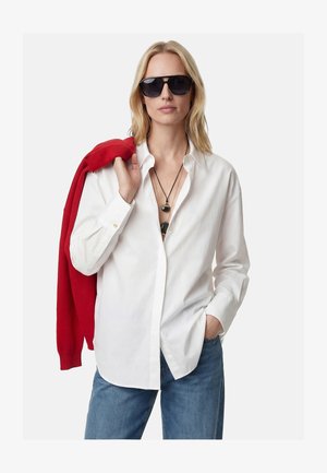 Donna bionda con occhiali da sole neri, camicia bianca con bottoni, jeans blu, collane a strati, tiene un maglione rosso sulla spalla, mano in tasca.