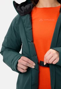 Groene gewatteerde jas met zwarte binnenkant, voorzien van een rits en drukknopsluiting. Draagt over een feloranje shirt met merklogo.