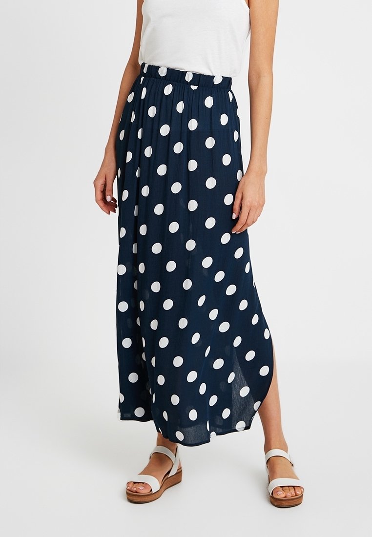 Femme portant une jupe bleu marine taille haute, longueur cheville, ornée de grands pois blancs, un haut sans manches blanc et des sandales blanches.