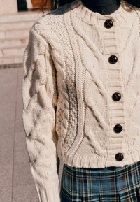 Cable-knit cream sweater with textured patterns, round neckline, and five contrasting black buttons. Paired with a plaid skirt.