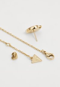 Gold jewelry set including a pair of stud earrings and a chain with a triangular pendant featuring the word "Guess." Smooth and shiny finish.