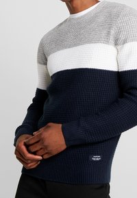 Man wearing a textured knit sweater with gray, white, and navy horizontal color blocks, holding hands in front of black pants.