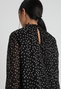 Schwarze, transparente Bluse mit kleinen Blumenmustern in Weiß und Pink, einem Schlüsselbein-Ausschnitt auf der Rückseite und einem rüschierten Halsausschnitt.