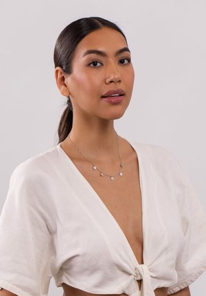 Silberne Kettenhalskette mit fünf kleinen, dekorativen Charms. Das Model trägt eine cremefarbene Bluse mit kurzen Ärmeln und lockerer Passform.